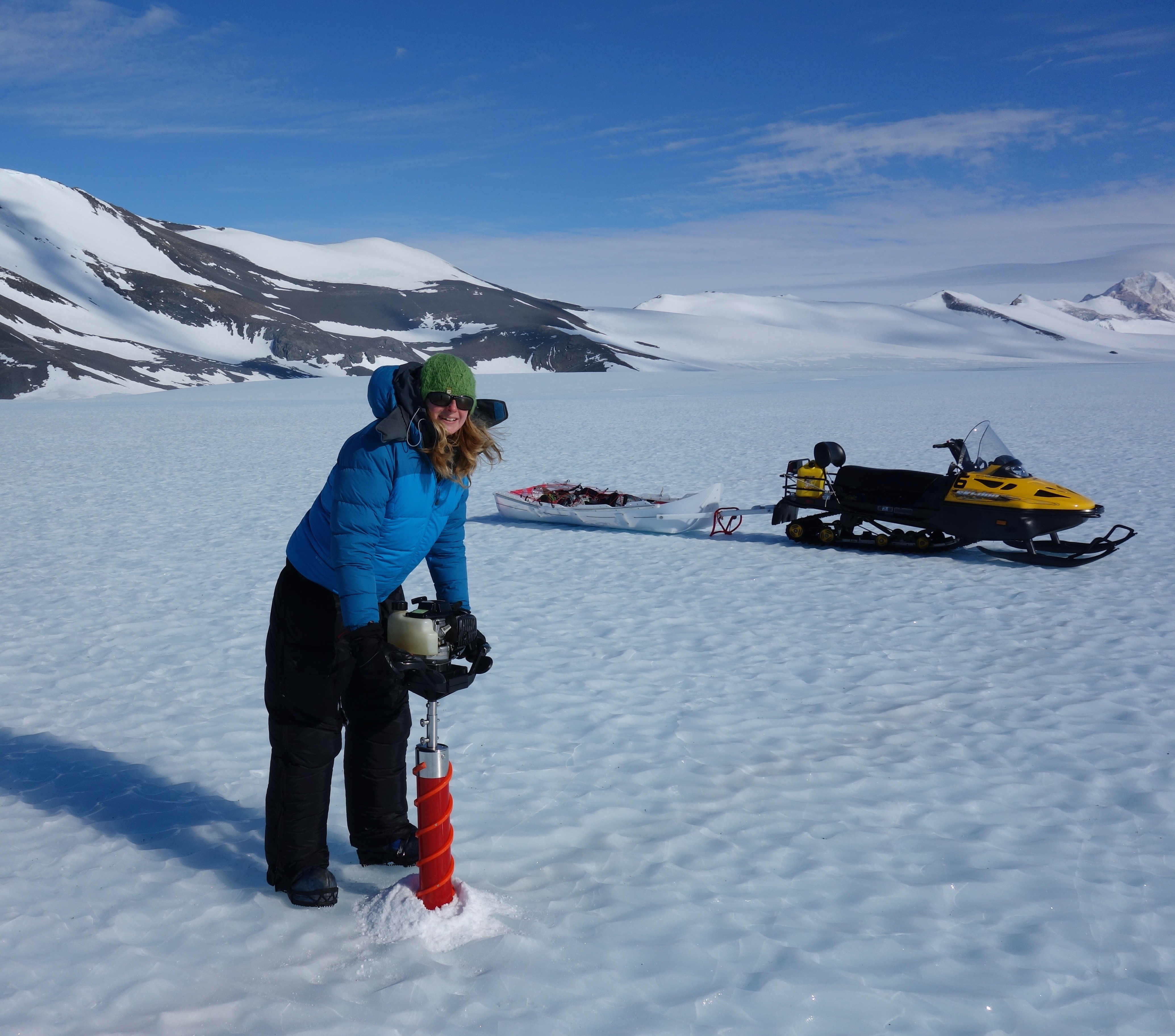 Glaciologist Dr Helen Millman drilling into ice with a long red drill. She is wearing black down trousers, a large blue down jacket, black gloves, a green woolly hat and sunglasses. There is a yellow skidoo with a white trailer in the background and some small mountains. The sky is bright blue.